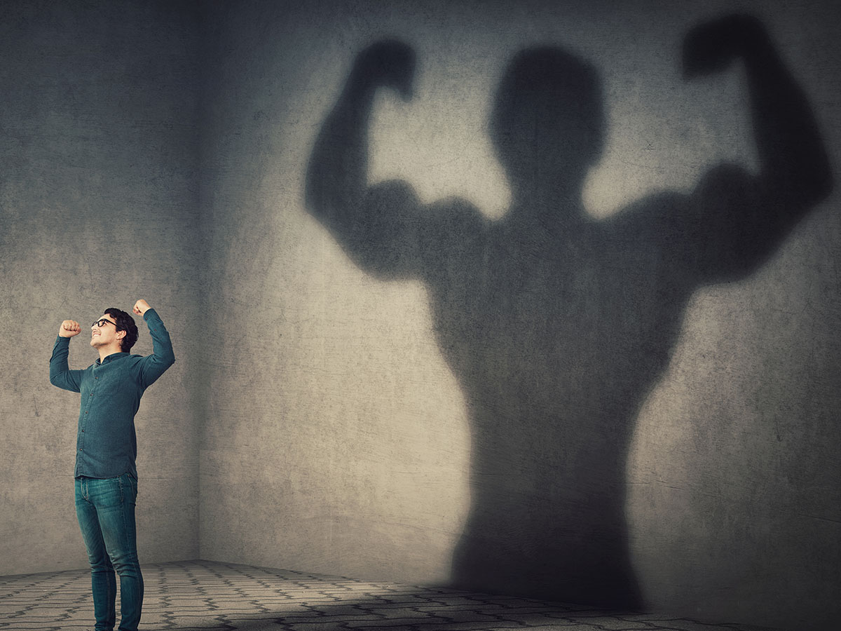 Man flexing in a grey room with his shadow also flexing but much more muscular.